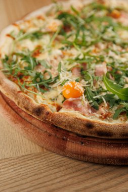 Yeşiller, peynir ve yumurta sarısı gibi çeşitli malzemelerden oluşan bir pizzaya yakından bakmak. Pizza, sıcak bir yemek ortamındaki ahşap servis tahtasına dayanır..