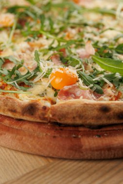 Jambonlu, peynirli, roka aromalı lezzetli bir pizza ve ahşap bir tahtanın üzerinde parlak yumurta sarısı servis edilir. Doğal ışık tabağı aydınlatırken, ortam sıradan..