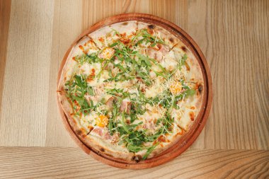 Yeni yapılmış bir pizza, jambon, peynir, roka ve portakal dilimlerini içeren malzemelerle gösterilir. Konforlu bir restoran ortamında ahşap bir tahtada sunuluyor..
