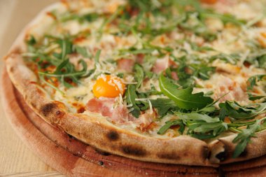 Jambon, roka ve yumurtalı taze pişmiş pizza servis edilmeye hazır ahşap bir tahtaya oturur. Bu yemek, sıradan bir yemek alanında İtalyan mutfağının lezzetlerini ön plana çıkarıyor..