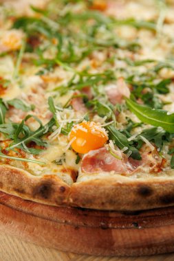 Taze bir pizza üzerine erimiş peynir, roka, jambon parçaları ve ortada çiğ yumurta sarısı. Tabak ahşap bir tahtada servis edilir, keyif verir..