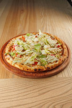 Marul, domates ve dolma biberli taze pizza ahşap bir tepsiye konur. Sahne, ahşap detaylarla dolu basit bir yemek alanını gösteriyor..
