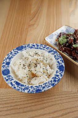 Beyaz pirinç, koyu baharatlı mavi desenli bir kasede sunulur. Lezzetli bir et tabağı, ahşap bir masada, pirincin yanında oturmuş, tadını çıkarmaya hazır..