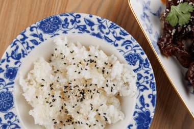 Asya mutfağından lezzetli bir tabağın yanına siyah susam tohumları serpiştirilmiş beyaz pirinçle dolu bir kase. Kurulumda ahşap bir masa var..