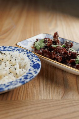 Çeşitli besin maddeleri ahşap bir masada, solda pirinç ve sağda şifalı otlarla dolu bir tabak et bulunuyor. Yemek, Ukrayna ve Japon mutfağından lezzetler sunuyor..