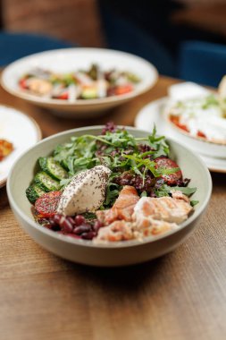 Farklı yemek kaseleri ahşap bir masada yeşil fasulye ve kavrulmuş sebzelerin yer aldığı bir salata üzerine kuruludur. Arka planda diğer yemekler mevcuttur.