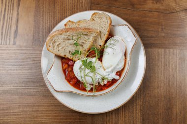 İki haşlanmış yumurta, taze yeşilliklerle süslenmiş, sıcak bir kafede basit bir tabakta servis edilmiş, zengin bir domates yahnisinin üzerinde oturur..