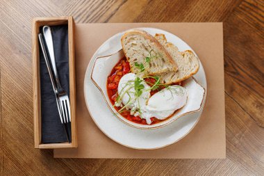 Bir kase haşlanmış yumurta, otlarla süslenmiş, ahşap bir masada servis edilmeye hazır, kabuklu ekmek dilimlerinin yanında, zengin domates sosunun içinde oturur..