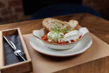 İki haşlanmış yumurta, basit bir tabakta taze ekmek dilimlerinin yanında zengin domates sosunun içinde oturur. Kurulumda sıcak bir ahşap masa ve minimalist çatal bıçak bulunuyor..