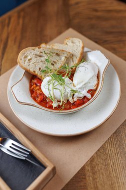 Haşlanmış yumurta, yanında bir dilim ekmek ile birlikte bir tabak sebze ve sosun üzerinde oturur. Sunumda bir çatal ve tahta masa yüzeyi yer alıyor..