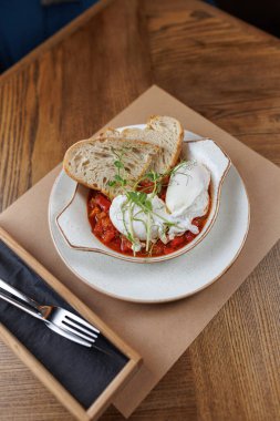 Bir tabak içinde bol domates sosunda dinlenen haşlanmış yumurtalar ve yanında bir dilim ekmek bulunur. Ortam akşam yemeği için mükemmel bir ortam sunuyor..