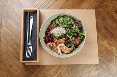 Izgara tavuk, taze salata, salatalık, salatalık, domates ve fasulye dolu bir kase, basit bir kabın içindeki çatal ve bıçağın yanındaki ahşap bir yüzeye yerleştirilir..