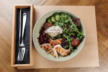 Ahşap bir masada ızgara tavuk, karışık yeşillikler, kiraz domatesleri, brokoli ve böbrek fasulyeleriyle dolu bir kase sunulur. Bir çatal kasenin yanında duruyor..