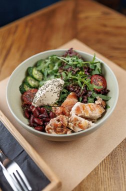 Sağlıklı bir kasede ızgara tavuk parçaları, kırmızı fasulyeler, kiraz domatesleri ve ahşap bir masada açık yeşil bir kasede servis edilen yeşiller yer alır..