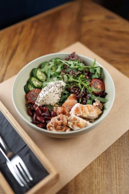 Izgara tavuk, kırmızı fasulye, dilimlenmiş sebzeler ve yoğurtla kaplı taze sebzelerle dolu bir kase restoran ortamında ahşap bir masada oturur..
