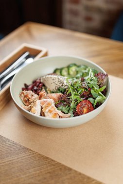 Bir kasede ahşap bir masada ızgara tavuk, karışık yeşillikler, salatalık ve kiraz domatesleri yer alıyor. Misafirler bu sıradan lokantada değişik tatlardan hoşlanırlar..