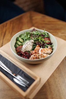 Taze sebzelerle dolu bir kase, ızgara tavuk, dilimlenmiş kabak ve fasulye bir restorandaki ahşap masada oturur. Basit bir ahşap tutacağa yakın bir çatal ve bıçak yerleştirilir..