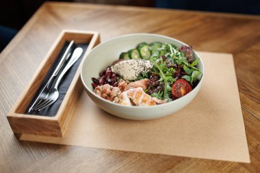 Bir kase ızgara somon, taze karışık sebze, kırmızı fasulye, dilimlenmiş salatalık ve vişneli domatesle doludur. Çatal bıçak takımı, sıradan bir yemek yerinde, ahşap bir masanın üzerine konur..