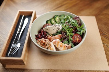 Taze yeşillik, ızgara tavuk, kiraz domatesleri, salatalık ve kafedeki ahşap bir masada duran yoğurt dolu bir kase. Çatal ve bıçak kâsenin yanında..