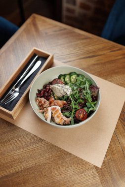 Taze salata, ızgara tavuk, salatalık, kiraz domatesleri ve otlarla dolu bir kase ahşap bir masada oturur. Çatal ve bıçaklarla dolu ahşap bir çatal bıçak tutacağı var..