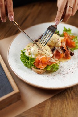 Bir insan somon yumurtalarını beyaz bir tabakta marulla füme olarak kesmek için çatal ve bıçak kullanır. Dekor, ahşap mobilyaları olan sıcak bir kafe..