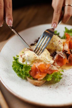 Bir insan, içinde füme somon ve taze yeşillik bulunan bir tabak yumurtayı kesiyor. Tabak, hareketli bir restoranda yuvarlak bir tabakta servis edilir. Atmosfer canlı ve sıcak..