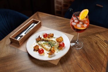 Güzel bir kaplama balık tabağı ahşap bir masada oturur. Kiraz domatesleri yemeğe eşlik ederken, portakal dilimiyle süslenmiş bir içecek de renk ve tat katıyor..