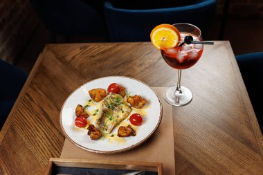 Kiraz domatesleri ve çıtır çıtır patateslerle birlikte bir tabak balık, portakalla süslenmiş canlı bir içeceğin yanında, ahşap bir masada oturuyor..