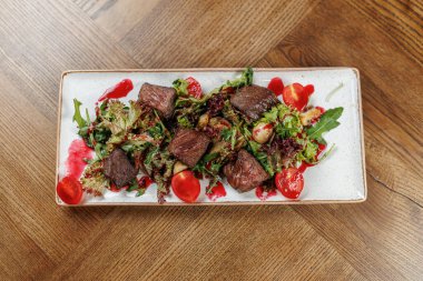 Izgara sığır eti parçaları karışık salata yeşillikleri üzerine kaplanmış, kiraz domatesleri ve renkli bir sosla süslenmiş. Bu yemek çeşitli aromalar ve sunumlar sunar.