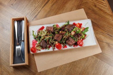 Izgara et parçaları beyaz bir tabakta karışık yeşillik ve vişneli domateslerle sunuluyor. Bu yemek, bir restoranda, ahşap bir masada servis edilir..