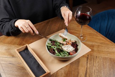Bir kişi ahşap bir yemek masasında oturuyor, tavuk ve sebzelerle taze bir salatayı kesiyor. Kasenin yanında bir kadeh kırmızı şarap var..