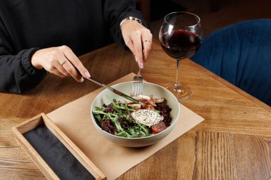 Bir insan sebze ve domatesten yapılmış bir salatada bir parça tavuk kesiyor. Sıcak bir restoranın atmosferinde masanın yanında bir bardak kırmızı şarap duruyor..