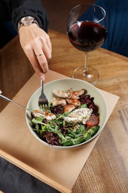 Bir el çatal tutar, taze yeşil salatayı ızgara tavuk, fasulye ve sebzeyle karıştırır. Gün batımından sonra sıcak ve davetkar bir restoranın yanında bir kadeh kırmızı şarap duruyor..
