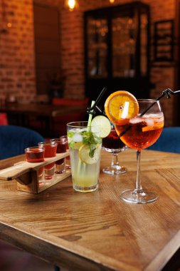 İçkiler bir restorandaki ahşap bir masada servis edilir. Naneli, limonlu, salatalıklı ve buzlu uzun bir bardak portakallı süslü bir kokteylin yanında duruyor. Çekim tepsisi görünür..