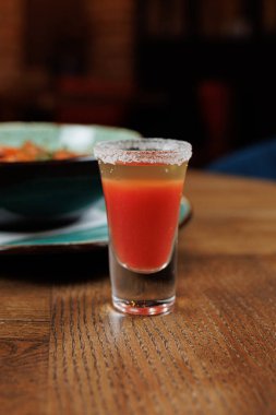 Renkli bir içecek, bir kase yemeğin yanındaki ahşap masada bir bardakta oturur. İçkinin tuzlu bir kenarı ve canlı bir tonu var. Canlı bir yemek atmosferinde tatmaya davet edici bir renk..