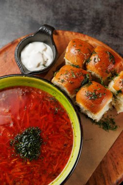 Bir kase canlı kırmızı sebzeli borş yumuşak ekmek rulolarının yanında oturur. Yanında küçük bir tabak kremalı sos var, güzel bir yemek sunumu yaratıyor..