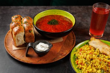 Otlu bir kase kırmızı çorba üç ekmek rulosunun yanında ahşap bir tabakta oturur. Pirinç ve içecek sıradan bir yemek alanında yemeği tamamlar..