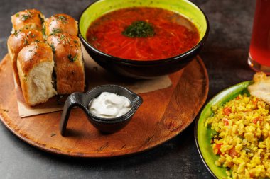 Bir kase çorba, yumuşak ekmek ruloları, küçük bir tabak ekşi krema ve renkli bir tabak pirinç de dahil olmak üzere çeşitli yiyecekler ahşap bir yüzeyde sergilenmektedir..