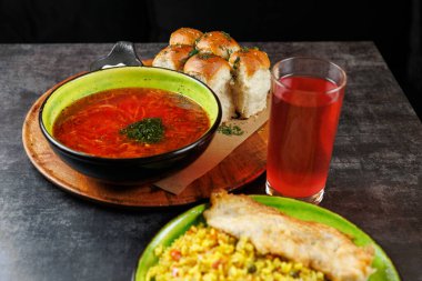 Masada bir kase borş çorbası, ekmek ruloları, bir bardak içecek ve bir tabak pirinçli balık yer alıyor. Yemekler, ortak bir yemek deneyimi için kültürel yemekler karışımını gösteriyor..