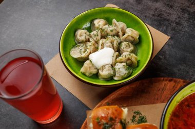 Ekşi krema ve otlarla kaplı yeşil bir tabakta bir kase Ukrayna hamur köftesi duruyor. Bir bardak kırmızı içecek, ahşap bir masanın üzerine konur ve geleneksel yemek sunumunu sunar..