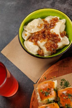 Üzerine tatlı karamelli soğan konmuş hamur köfteleri kahverengi bir masada serinletici bir içeceğin yanında yeşil bir kasede oturur. Arkaplanı çeşitli yemekler doldurur..