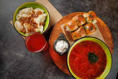 Parlak kâselerde soğanlı hamur tatlısı, bitkilerle süslenmiş canlı kırmızı bir çorba, fırında rulo ekmek ve yanında ekşi krema. Serinletici içecek yemeği tamamlar..