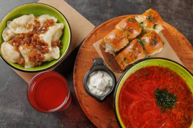 Bir kase kırmızı borş, üzerinde et olan hamur köftelerinin yanında oturur. Sarımsaklı ekmek, ekşi krema ve içecek ile birlikte ahşap bir tabakta mevcuttur..