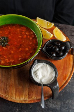 Bir kase borş, yanında limon dilimleri, zeytin ve ekşi krema olan ahşap bir tepside oturur. Taze otlar çorbayı ekstra lezzet için süslüyor..