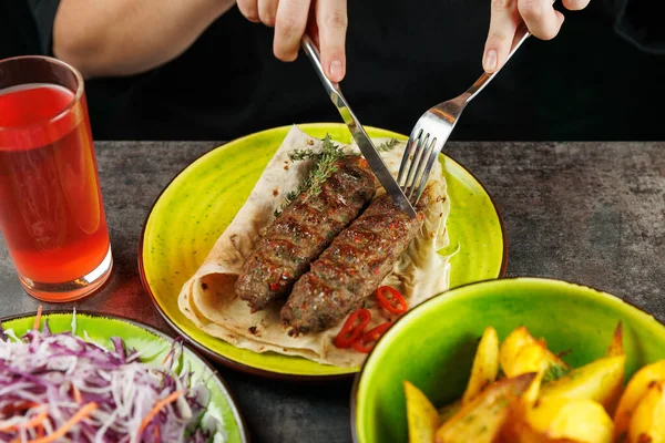 Biri salata ve içecekle servis edilen bir tabakta iki parça şiş et kesiyor. Dekor canlı renkler ve taze gıda seçenekleriyle dolu..