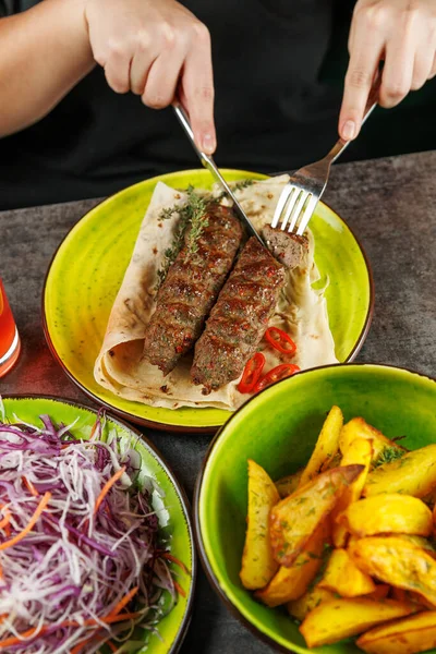 Renkli bir sebze salatası ve canlı bir masa üzerinde altın-kahverengi patates eşliğinde kızarmış kebap ile servis edilen yassı ekmeğin üzerinde ızgara kebap kesen bir insan..
