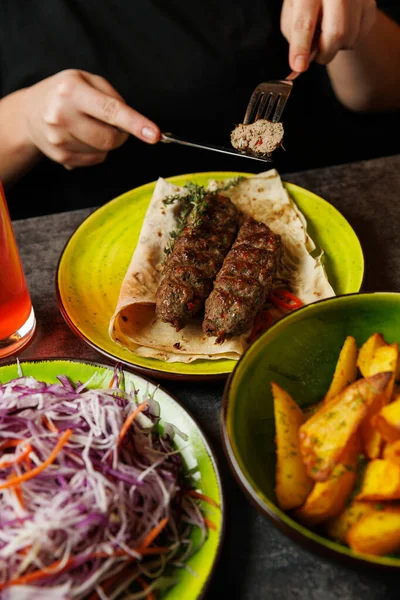 Bir lokanta, tortilla üzerinde ızgara kebaplara baharat eklemek için çatal kullanıyor. Yakınlarda, renkli tabaklar taze salata ve kızarmış patates var. Bir içki yemeği tamamlar..