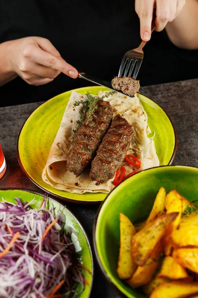 Bir insan ekmek üzerine ızgara et servis etmek için çatal kullanıyor. Masanın üzerinde renkli tabakların yanında salata ve kızarmış patates var. Kurgu, zengin bir aşçılık deneyimini yakalıyor..