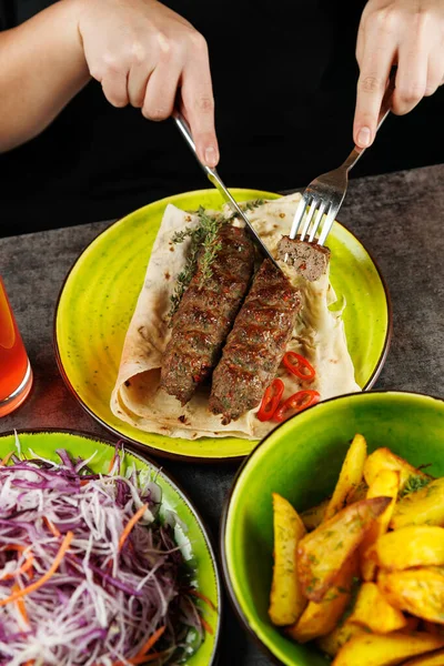 Bir insan ekmek üzerine ızgara kebap kesmek için çatal ve bıçak kullanır. Yakınlarda patates dilimleri ve taze bir salata var. Tatmin edici bir yemek hazırlıyorlar..