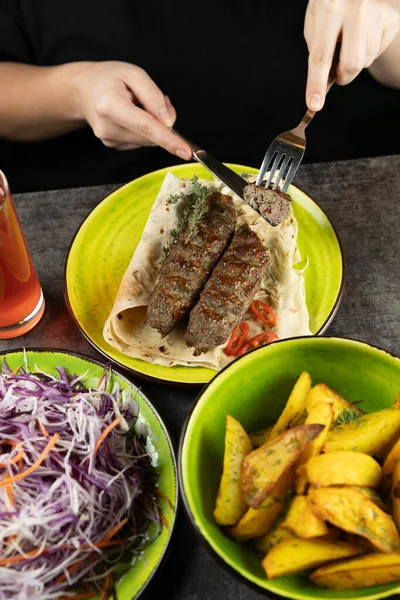 Bir insan, yanında kızarmış patates ve taze salatayla birlikte kızarmış kebaplı bir yemeğin tadını çıkarıyor. Renkli bir içecek yemek deneyimine katkı sağlar..